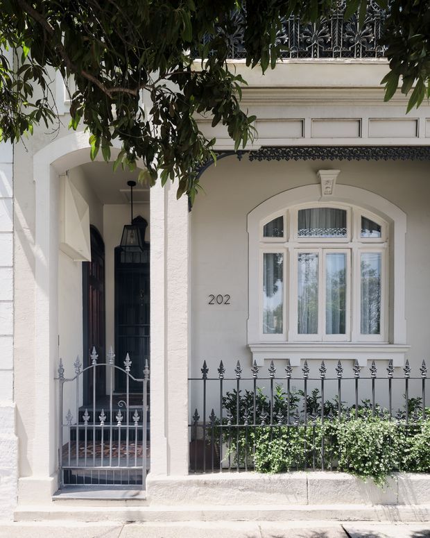 Street-level view of a refined historic townhouse facade with a recessed entry porch, arched white-trimmed bay window, ornate wrought-iron balcony and front fencing, and a pale rendered exterior. Mature greenery partially frames the upper facade, with the number 202 visible beside the entrance.