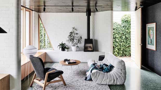 Bright contemporary living room with white-painted brick walls, extensive timber-framed glazing, a white sculptural sofa, dark upholstered lounge chair, timber coffee table, indoor plants, and a compact black fireplace set beneath a central structural post. The room has a distinctive timber-lined ceiling with exposed track lighting, polished pale flooring, and a dark textured feature wall with framed artwork.