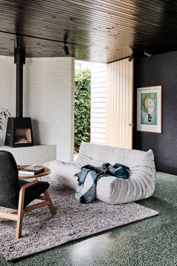 A stylish living room with a low wood-slatted ceiling, white-painted brick fireplace surround, black wood-burning fireplace, textured dark feature wall, light timber panelled door, white sculptural sofa, dark lounge chair, patterned rug, and green terrazzo-like flooring. The room opens toward a leafy exterior courtyard or garden, with framed artwork and track lighting adding contemporary character.