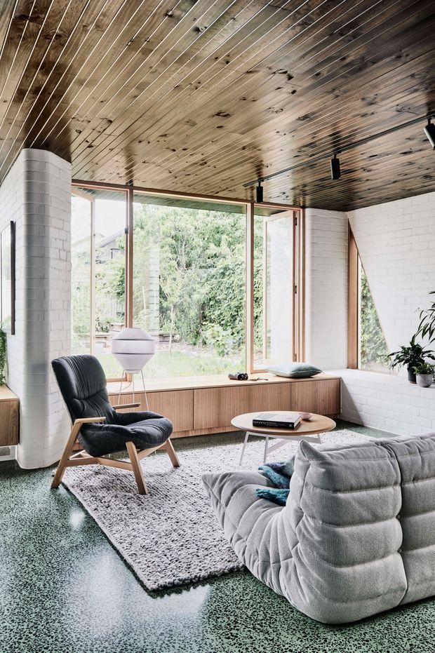 A light-filled living room with floor-to-ceiling timber-framed glazing opening to a leafy garden. The room features a white-painted brick fireplace or structural column, a dark timber-lined ceiling with exposed boards, terrazzo-style green flooring, a textured neutral rug, a low timber media console, a sculptural lounge chair, a rounded coffee table, and a large plush modular sofa. The interior has a refined mid-century and contemporary character with strong natural materials and indoor-outdoor connection.