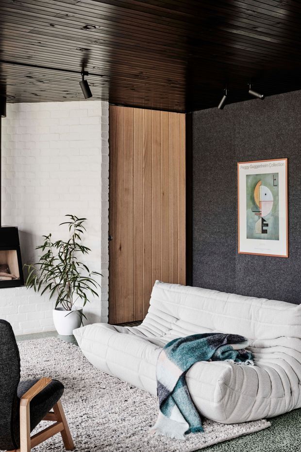 A cozy, design-focused living room featuring a white quilted sofa, textured pale rug, black leather lounge chair, indoor plant, framed artwork, and a dark timber-lined ceiling. The room combines a white painted brick wall, vertical timber wall panelling, and a dark charcoal acoustic-looking feature wall with directional track lighting.