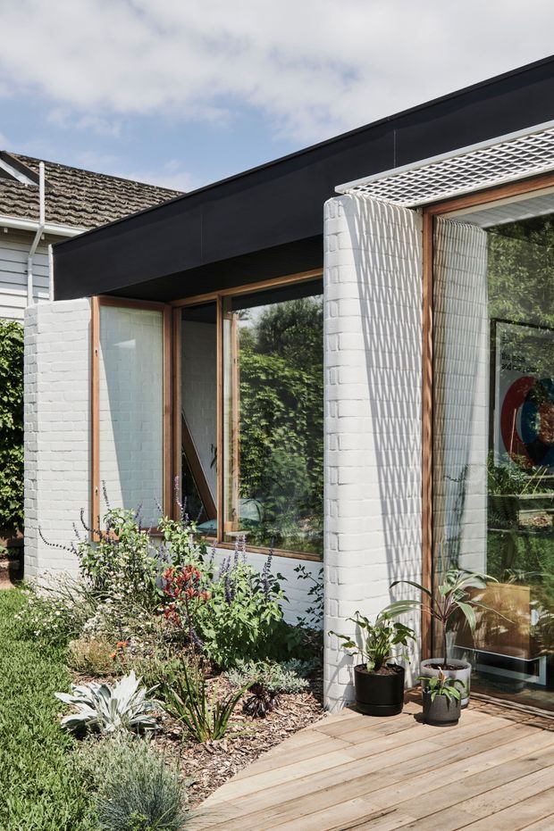 Exterior view of a contemporary residential extension with a low black roofline, white-painted brick walls, timber-framed glazing, and a timber deck. The facade incorporates a white lattice-style sunshade over a large window, with a landscaped garden and potted plants in the foreground. An older weatherboard and tiled-roof structure is visible behind the extension.