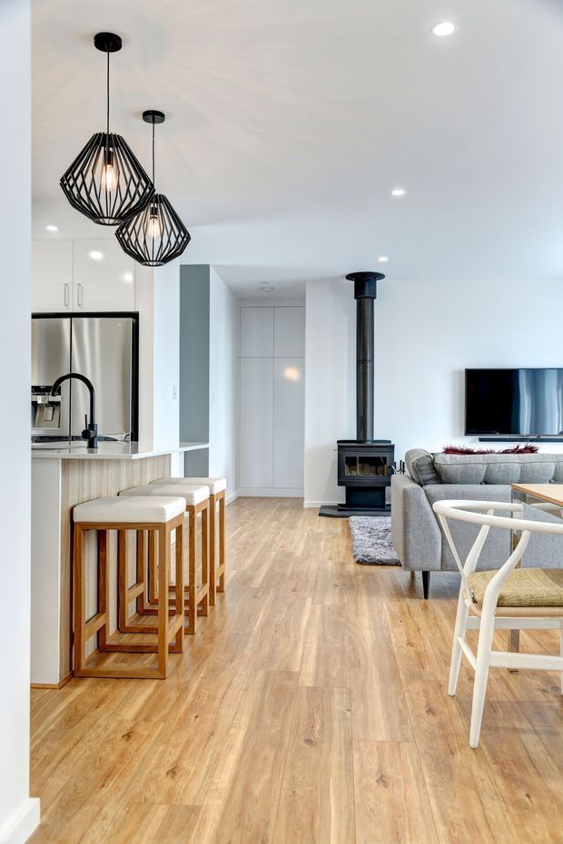 Bright open-plan interior combining a compact kitchen, island seating, and living area. The space has white walls and cabinetry, light timber-look flooring, stainless-steel appliances, a black freestanding wood-burning stove, grey upholstered seating, geometric black pendant lights, and contemporary dining chairs. The kitchen and living room are visually connected in a clean, airy layout.