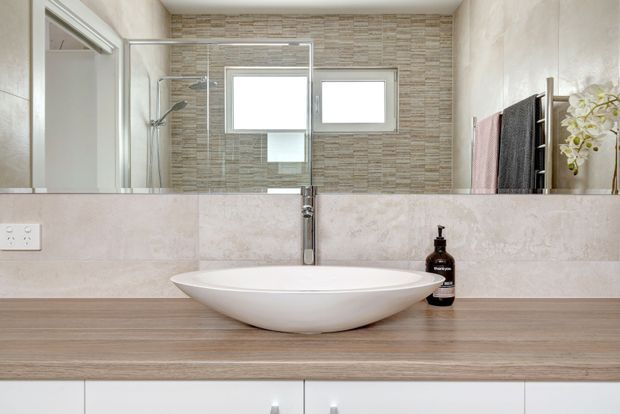 A bright contemporary bathroom vanity with a white vessel basin set on a timber-look benchtop, pale stone-look wall tiles, a large mirror, and a glass-enclosed shower visible in reflection. The room includes a wall-mounted tap, small horizontal window, towel rail, and simple white cabinetry.