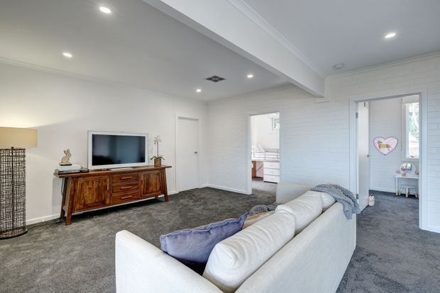 Bright open-plan living room with a large white sectional sofa, grey carpet, white painted brick walls, recessed ceiling lights, and a timber sideboard supporting a television. Open doorways lead to adjoining rooms, including a partially visible bedroom or dressing area and a small child's room.