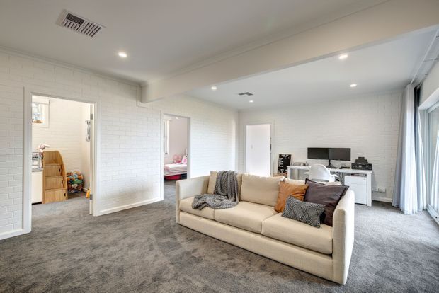 Bright, spacious living room with a large beige sectional sofa, grey carpet, white painted brick walls, recessed ceiling lights, and broad glazing with curtains. Open doorways connect to adjacent rooms, while a wall-mounted TV and low media unit occupy the rear wall.