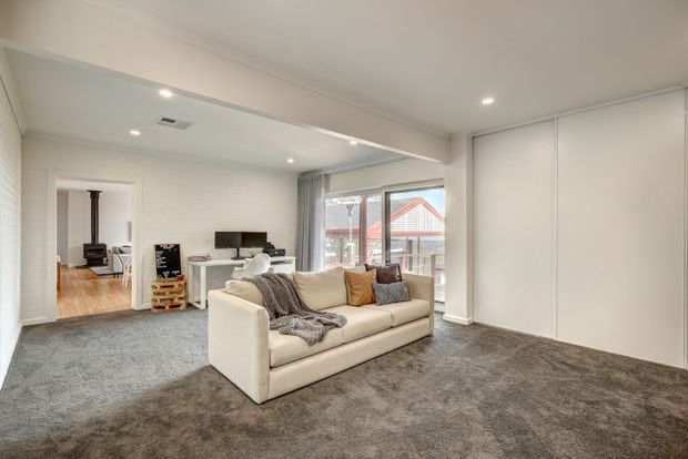 Bright, spacious contemporary living room with a large cream sectional sofa, grey carpet, white walls and built-in full-height cabinetry. Recessed ceiling lights, wide glazing with curtains, and an adjoining dining area and fireplace room are visible.