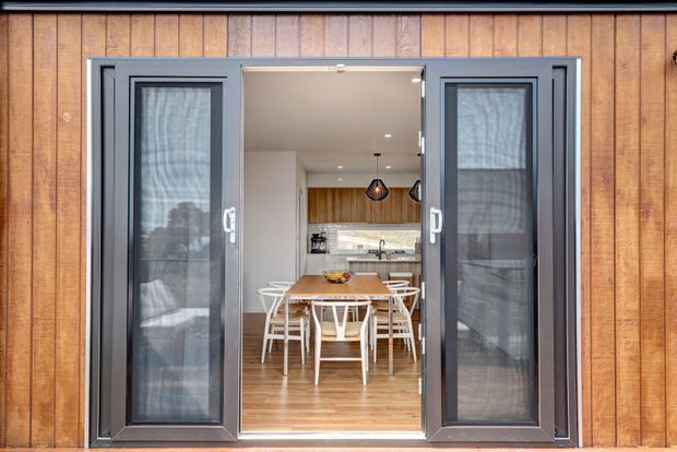 View from outside through open dark-framed glazed folding doors into a bright dining and kitchen area. The exterior is clad in warm vertical timber boards, while the interior features timber flooring, a wood dining table with white chairs, white walls, and a compact kitchen with pendant lighting.