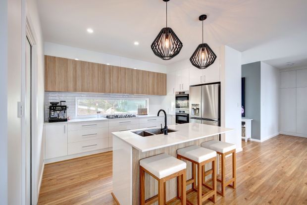 Bright contemporary open-plan kitchen with white cabinetry, a large white island and integrated breakfast bar, warm timber flooring, a timber overhead cabinet feature, stainless-steel appliances, and two black geometric pendant lights. The kitchen opens toward a hallway or adjacent living area.