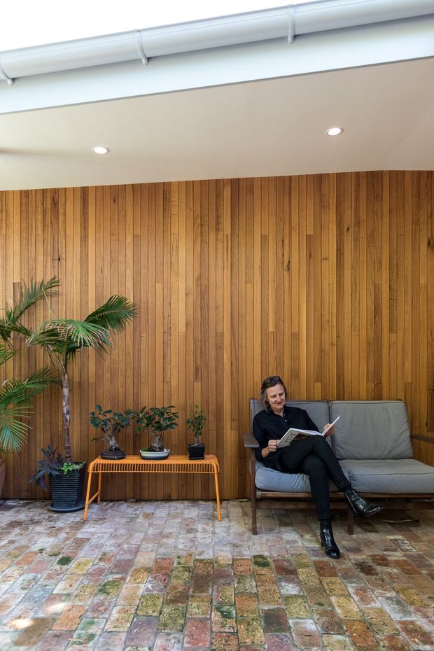 A stylish indoor seating area with a full-height vertical timber feature wall, exposed white ceiling services, recessed downlights, and a reclaimed brick floor. A grey upholstered sofa sits beside a slim orange-toned bench displaying potted plants, while a person reads on the sofa. The space has a warm, relaxed, plant-filled character with an industrial-influenced fit-out.
