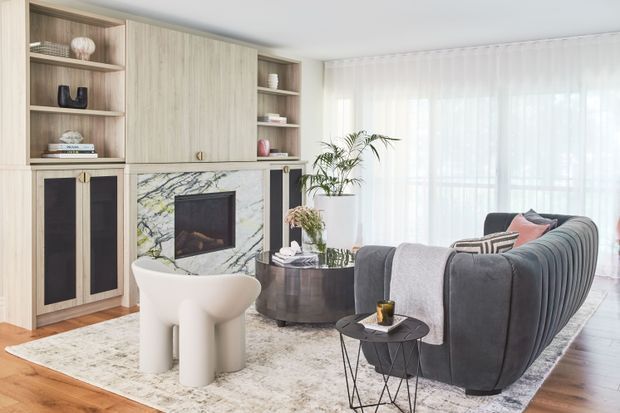 Bright contemporary living room with a sculptural white lounge chair, a large curved dark-grey sofa, round nesting side tables, and a marble-faced built-in fireplace wall with integrated timber cabinetry and open shelving. Full-height sheer curtains diffuse daylight, while a large rug, indoor plant, and restrained decor create a refined, comfortable setting.