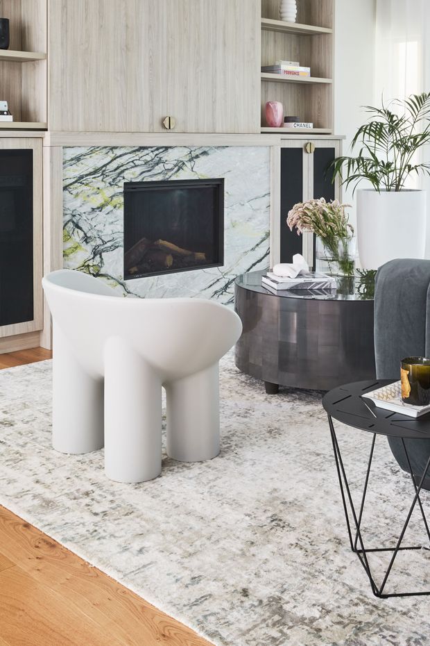 A refined contemporary living room centred on a built-in fireplace with a dramatic veined stone surround and integrated light-wood cabinetry. The room includes a sculptural white armchair, dark rounded coffee table, charcoal upholstered seating, a black geometric side table, a large muted patterned rug, and warm timber flooring. Neutral materials and soft natural light create a polished, inviting interior.
