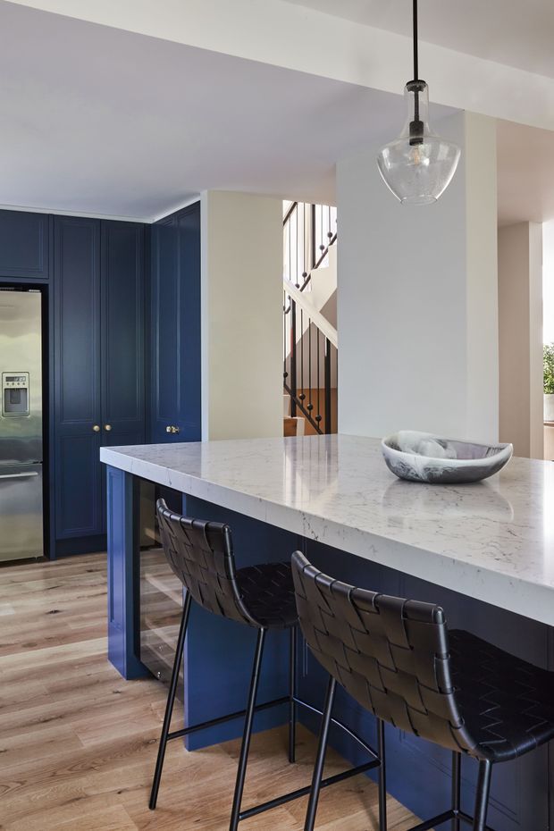 A refined kitchen interior with a large white marble-look island, deep navy-blue shaker cabinetry, integrated stainless-steel refrigerator, timber flooring, and two dark woven bar stools. A clear glass pendant light hangs above the island, while a staircase is visible beyond the kitchen.