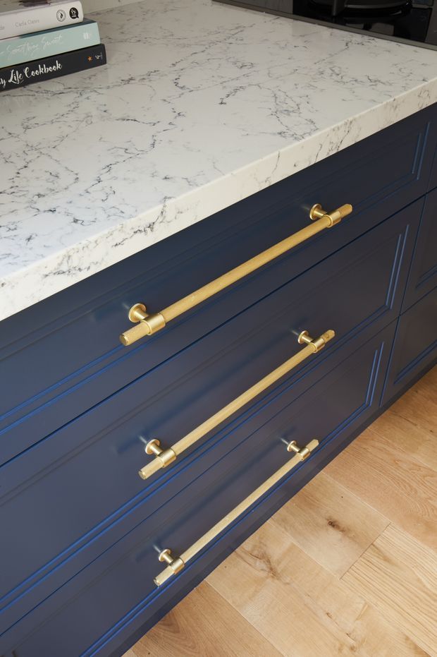 Close-up view of a dark navy-blue kitchen island or cabinetry with recessed panel drawer fronts and long brushed brass-gold bar pulls. The island is topped with a white marble-look engineered stone benchtop featuring prominent grey veining, and sits over light natural timber flooring. A small stack of cookbooks is visible on the benchtop.