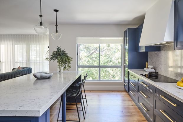 A bright contemporary kitchen viewed toward a large window, with a long white stone island, dark blue fixed cabinetry, brass-toned hardware, a marble-look splashback, pendant lights, timber flooring, and bar seating. An adjoining living area is partially visible at left.