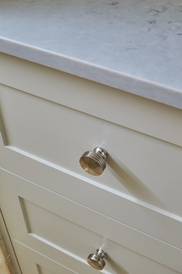 Close-up detail of cream painted shaker-style cabinetry with recessed drawer fronts and polished metallic round knobs, beneath a pale veined stone benchtop.