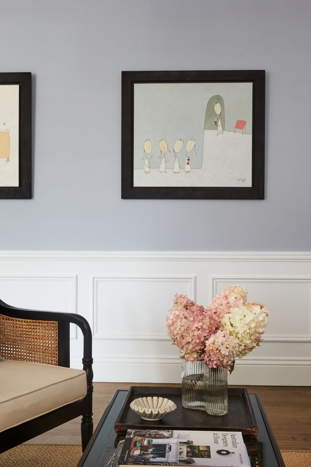 A refined living room vignette with pale grey walls, white decorative wall panelling, dark timber flooring, and framed artwork. A black cane-backed armchair sits beside a dark square coffee table holding a ribbed glass vase filled with soft pink and cream hydrangeas, a small decorative bowl, and a magazine. The composition has a classic, elegant residential interior feel with restrained neutral tones and floral accents.