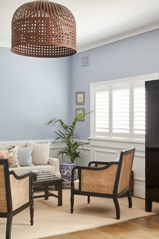 A refined living room with pale blue walls, white trim and plantation shutters, furnished with a cream sofa, black-framed cane armchairs, a dark wood coffee table, and a decorative blue-and-white planter. A large woven rattan pendant light hangs overhead, while a neutral textured rug sits on warm timber flooring. The room has a calm, coastal-traditional character with strong natural-material accents.
