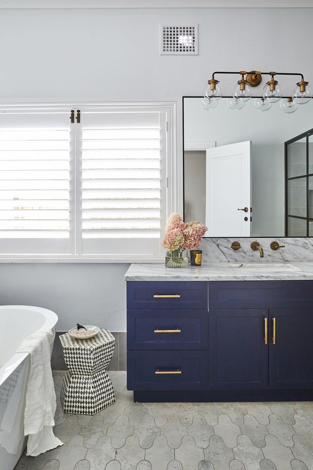 Bright, refined bathroom featuring a deep navy-blue vanity with brass hardware, a white and grey marble benchtop and integrated backsplash, wall-mounted brass fixtures, a large black-framed mirror, white plantation shutters, and a freestanding white bathtub. The floor is finished with patterned pale grey tiles, while a black-and-white geometric stool adds contrast.
