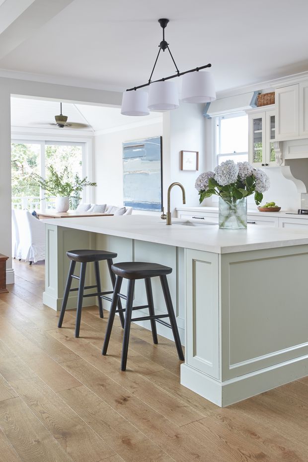 Bright, refined kitchen interior featuring a large pale sage-green island with white benchtops, paneled cabinetry, brass gooseneck tapware, dark wood bar stools, and a trio of pendant lights. Wide timber flooring connects to a light-filled living or sunroom area, with white cabinetry, soft blue artwork, and hydrangeas adding coastal character.