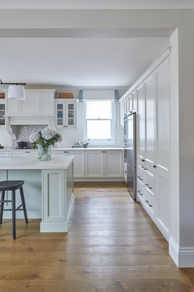 A bright, refined traditional kitchen with extensive white shaker-style cabinetry, a large pale island with panelled sides, integrated appliances, a decorative rangehood surround, and a patterned tile backsplash. Warm natural timber flooring contrasts with the cool white joinery, while a window with a pale blue Roman blind provides soft daylight. The kitchen includes a small dark stool and a central floral arrangement.