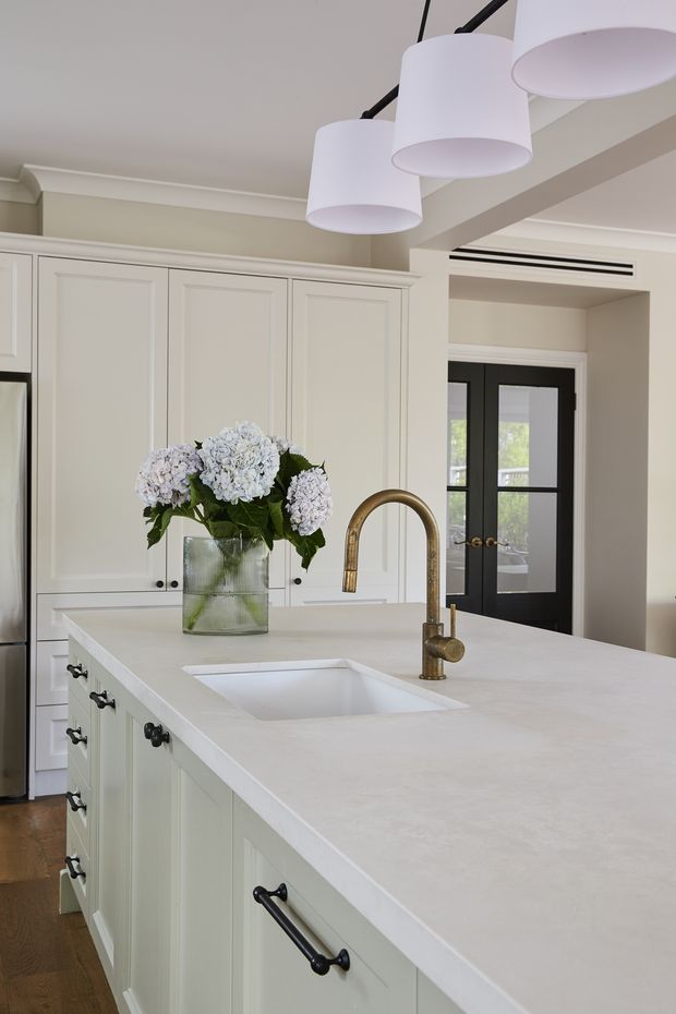 Bright, refined kitchen interior featuring a large white island with an integrated sink and aged brass gooseneck tap, pale paneled cabinetry with dark hardware, white pendant lights, and a black-framed glazed doorway. A vase of hydrangeas provides a soft decorative focal point.