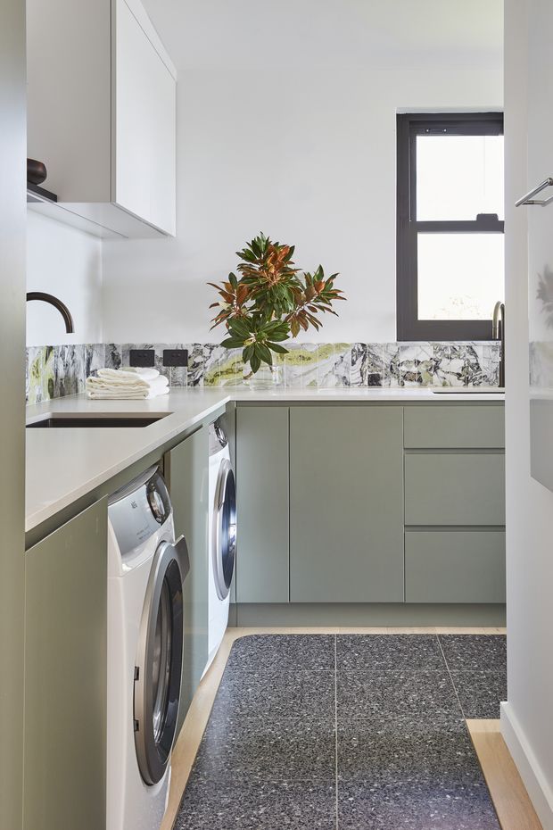 A compact, contemporary laundry room with muted green-grey built-in cabinetry, white benchtops, integrated front-loading washing machines, and a dramatic veined stone or marble-look splashback. A small window provides natural light, while dark grey terrazzo-style floor tiles and a leafy plant add contrast and texture.