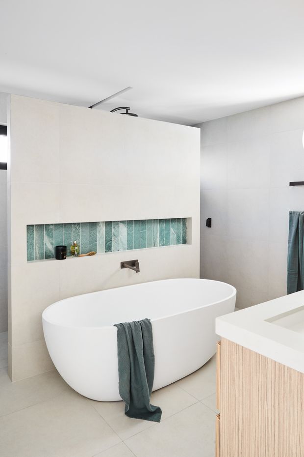 Bright contemporary bathroom featuring a freestanding white bathtub, wall-mounted matte dark tapware, pale large-format tiled walls and floor, and a recessed decorative niche lined with vertical turquoise tiles. A light timber vanity with a white benchtop is visible in the foreground.