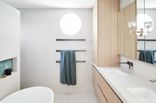 Bright contemporary bathroom with a freestanding white bathtub, double vanity, pale timber cabinetry, white solid-surface benchtop, large mirror, round wall opening or window, and dark horizontal towel rails. A small teal tile feature is visible beside the bath, while globe pendant lights add a refined accent.
