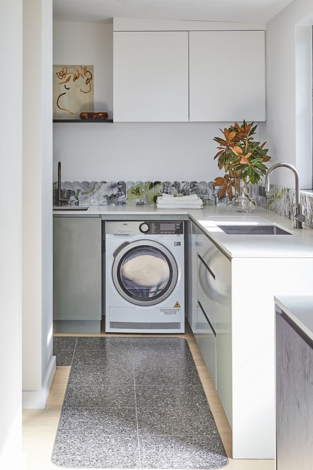 Bright, compact contemporary laundry with a U-shaped layout. White upper and lower cabinetry surrounds a front-loading washing machine, with a white benchtop, undermount sink and chrome gooseneck tap. A patterned grey and green tiled splashback adds visual interest, while dark terrazzo-look floor tiles and a runner ground the space. Natural light enters from the right, with a plant and small decorative objects providing warmth.
