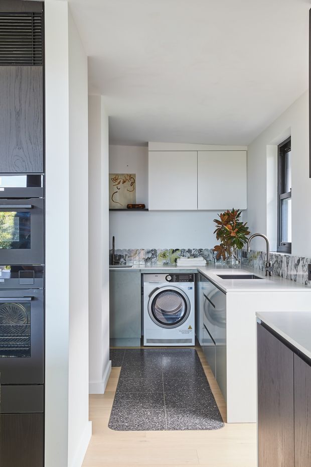 A bright, compact contemporary utility and laundry area viewed from an adjacent kitchen. The space features white upper and lower cabinetry, a front-loading washing machine, a stainless-steel sink with a high curved tap, a patterned grey-and-white splashback, dark terrazzo-like floor tiles, pale timber flooring outside the laundry zone, and dark-framed windows. Integrated dark cabinetry and appliances are visible along the left edge.