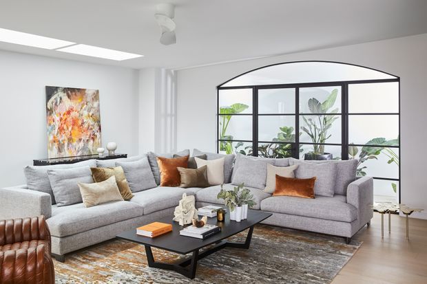 Bright contemporary living room featuring a large grey sectional sofa, black rectangular coffee table, layered patterned rug, and warm neutral accent cushions. A dramatic black-framed arched internal glass partition overlooks leafy planting, while an abstract artwork and sculptural decor add visual interest. The room has a white ceiling with recessed linear skylight-like lighting and pale timber flooring.