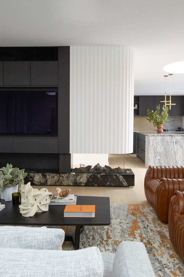 A refined contemporary living room with a dark built-in media wall, sculptural fluted white feature partition, and a linear stone-clad fireplace hearth. The room opens toward a sophisticated kitchen with dark cabinetry, a large veined stone island, and pendant lighting. Rich brown ribbed leather seating, pale upholstered chairs, a black coffee table, decorative objects, and a muted patterned rug create a luxurious, warm-toned interior.