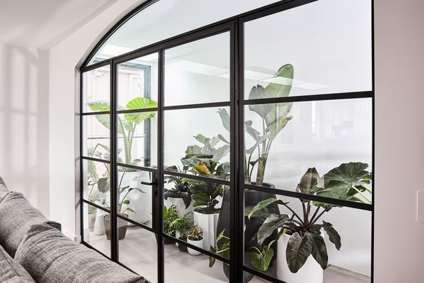 Bright contemporary interior with a black-framed, steel-look glazed partition featuring an arched upper section. The partition separates a light-filled indoor plant room or sunroom from a living area, with abundant potted tropical foliage visible behind the glass and part of a grey sofa in the foreground.