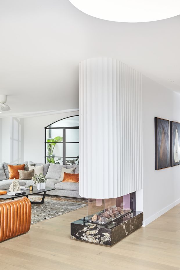 Bright contemporary living room featuring a sculptural curved fireplace divider with vertical white fluted detailing and a dark marble hearth. Pale timber flooring, a large grey sectional sofa with rust-orange cushions, black-framed internal glazing, artwork, and a coffee table create a refined open-plan interior. A large circular ceiling feature is visible above.