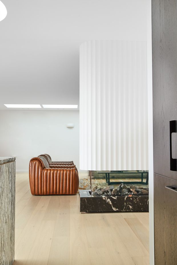 Bright contemporary living room viewed through a doorway, featuring pale timber flooring, a sculptural white fluted fireplace surround or room divider, a dark veined stone hearth, a low glass-and-metal console, and a distinctive caramel leather channel-tufted armchair. Minimal white walls and concealed linear lighting create a clean, refined interior.