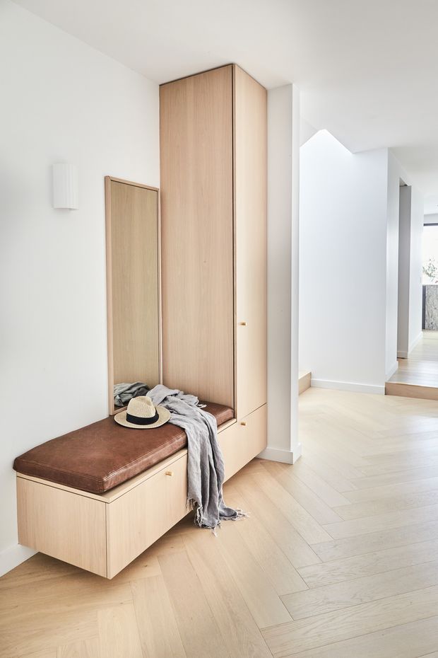 Bright minimalist residential entryway featuring a full-height light timber built-in cabinet with integrated bench seating, a brown leather cushion, mirror, and concealed storage. The space has white walls, pale timber flooring laid in a herringbone pattern, and a light, airy corridor extending beyond.