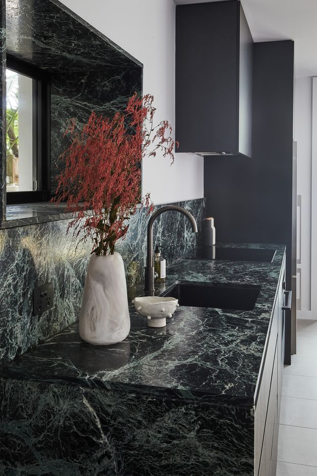 A refined contemporary kitchen focused on a dramatic dark green-black veined stone benchtop and matching splashback. The space includes matte charcoal cabinetry, a dark undermount sink with curved brushed-metal tapware, integrated appliances, and a sculptural white vase with red branches. A narrow window and light walls provide contrast, while pale flooring keeps the room bright.
