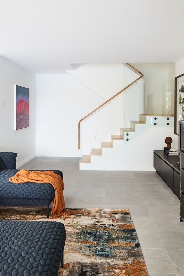 Bright contemporary bedroom and stair hall with a minimalist white interior. The room features a low dark upholstered bed with textured navy bedding, a vivid orange throw, a large patterned area rug, pale grey tiled flooring, white walls, and a sculptural open staircase with light timber treads, a slim timber handrail, and glass balustrades. Dark built-in cabinetry and framed artwork add contrast.