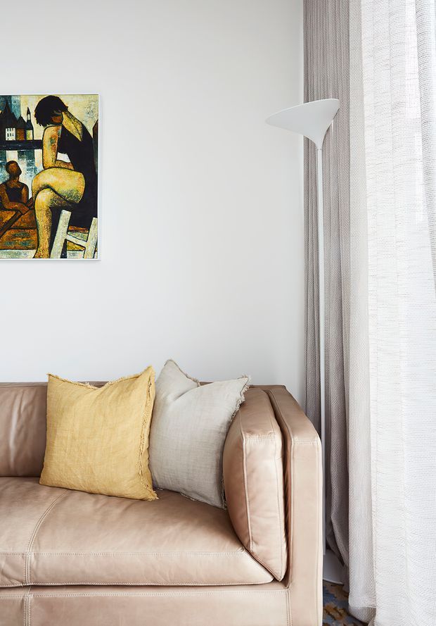 A softly lit living room corner featuring a blush-beige leather sofa with yellow and ivory cushions, a white sculptural floor lamp, sheer textured curtains, and a framed figurative artwork on a pale wall. The composition has a refined, warm, contemporary feel with subtle mid-century influence.