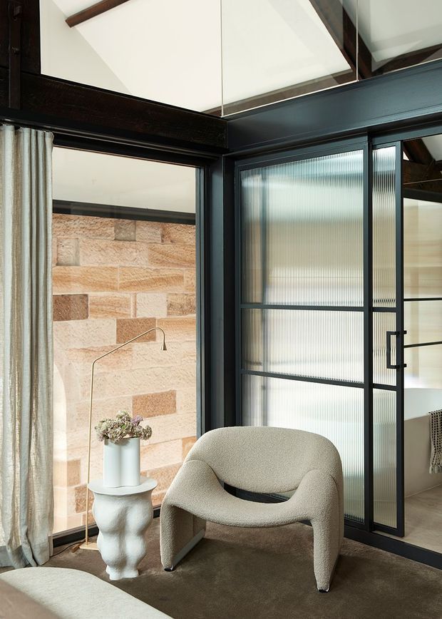 A refined interior corner with a sculptural cream boucle armchair, small white side table, and neutral curtains. Dark timber ceiling beams and black-framed ribbed translucent glass doors create a strong architectural contrast, while a warm sandstone block wall is visible beyond the glazing. The space has a calm, contemporary character with natural textures and carefully curated furniture.