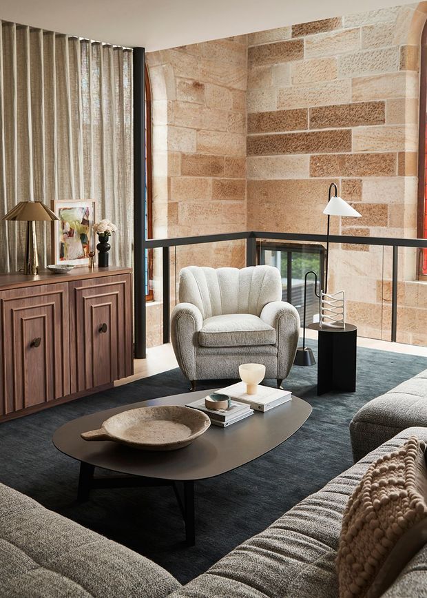 A warm, refined living room with a large upholstered armchair, sectional sofas, a dark sculptural coffee table, a timber sideboard, floor-to-ceiling curtains, and black-framed glazing. Textured tan stone masonry forms the feature wall and creates a strong architectural backdrop, while a glass balustrade overlooks a lower level.