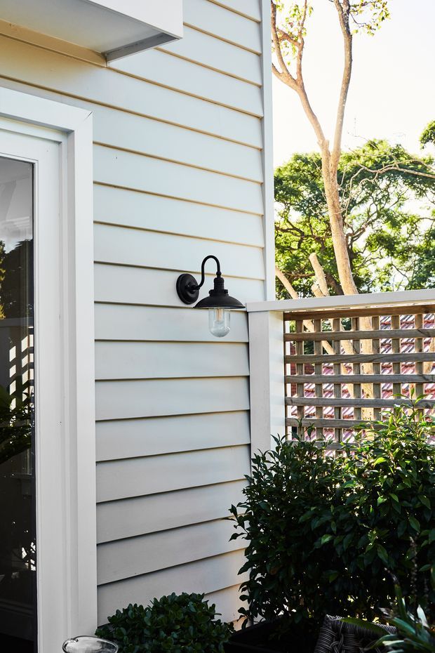 Close-up exterior view of a light-painted weatherboard-clad wall beside a glazed door, featuring a black wall-mounted outdoor lantern and a timber lattice privacy screen forming part of a small deck or terrace. Dense established planting is visible around the lower edge, with trees beyond.