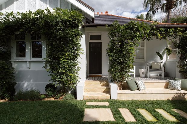 A leafy front garden leading to a small, elegant residential entry porch. The light-painted facade has white-framed windows, a dark timber front door, a tiled pitched roof, and a raised timber-look deck with built-in bench seating and cushions. Clipped hedges frame the entry, with stepping-stone paths crossing the lawn.