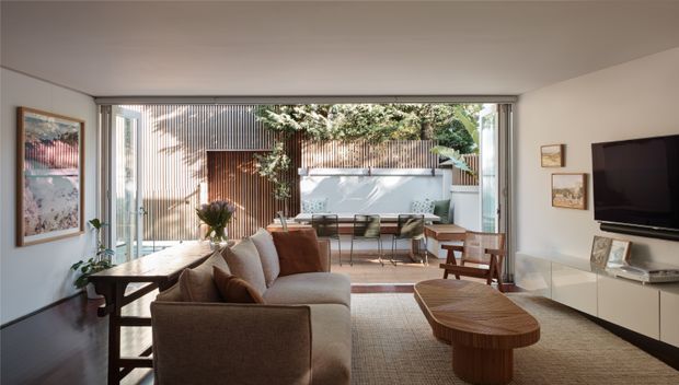 A bright, refined living room opening onto a landscaped courtyard through wide floor-to-ceiling glazed doors. The room features a neutral upholstered sofa, timber coffee table and dining table, built-in white media cabinetry, framed artwork, and warm timber screening and planting visible outside. The space has a contemporary indoor-outdoor connection and a calm, natural palette.