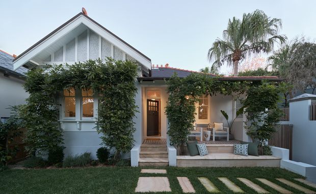 Front exterior of a single-storey cottage-style home with a prominent gabled facade, decorative white gable detailing, climbing greenery, a covered entry porch, and a paved stepping-stone path through the lawn. The facade is painted in pale grey and white, with a dark tiled roof and timber-framed doors and windows.