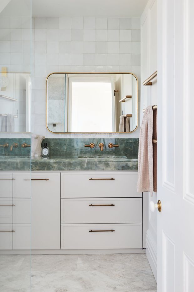 Bright, refined bathroom vanity with white tiled walls, pale stone-look flooring, white shaker-style cabinetry, and a green veined stone benchtop with integrated basin. A rounded-rectangle mirror with a warm metallic frame sits above brushed brass wall-mounted tapware. Brass hardware and a timber-toned towel rail add subtle warmth, while a partially open white paneled door frames the room.