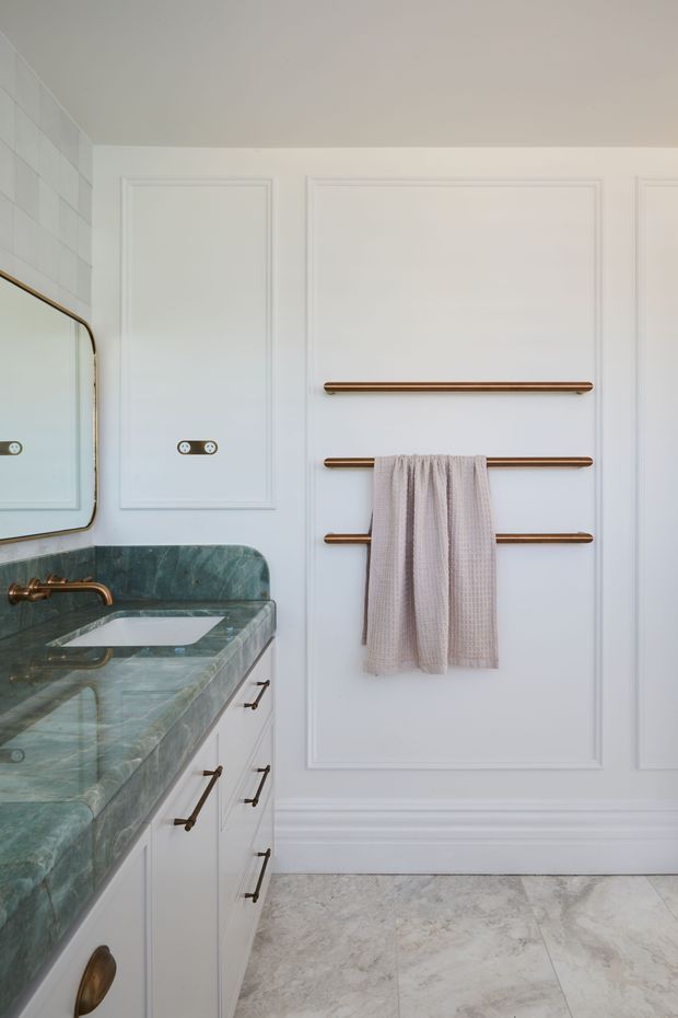 A refined bathroom vanity area with white decorative wall panelling, a green veined stone benchtop, integrated white basin, antique brass tapware and hardware, and three horizontal brass towel rails. The floor is finished in pale grey stone-look tile, and part of a rounded-edge mirror is visible.