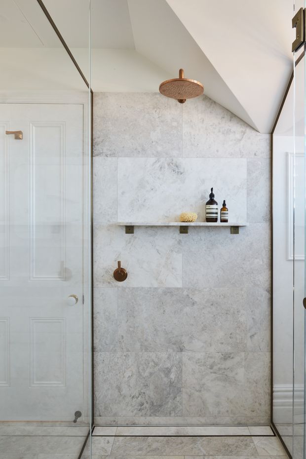 A bright, refined bathroom shower enclosure with full-height pale marble-look stone wall and floor tiles, a frameless glass screen, a recessed-style floating shelf, and warm copper-toned shower fittings. A white paneled door and traditional trim are visible beside the shower. The sloped ceiling adds an attic-like architectural detail.