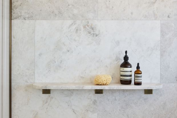 Close-up of a minimalist bathroom shower niche or wall shelf, featuring pale veined stone wall tiles and a matching floating stone ledge with dark brackets. Amber toiletry bottles and a natural sponge sit on the shelf.